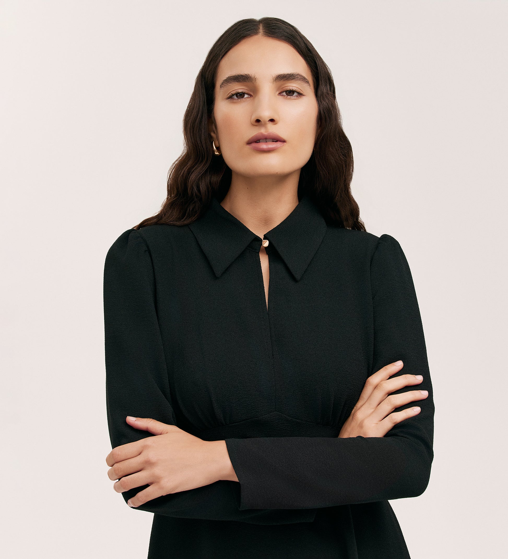 A person with long, wavy dark hair stands against a plain light background, arms crossed, wearing the Sabine Black Crepe Short Dress—a fitted mini dress in woven crepe with long sleeves.