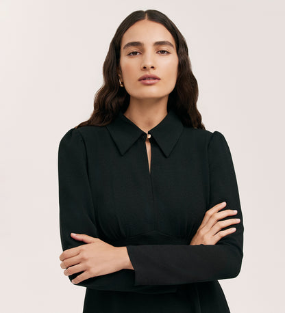 A person with long, wavy dark hair stands against a plain light background, arms crossed, wearing the Sabine Black Crepe Short Dress—a fitted mini dress in woven crepe with long sleeves.