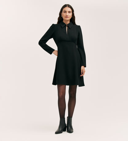 A woman with long dark hair and a neutral expression stands against a plain background wearing the Sabine Black Crepe Short Dress with long sleeves, a keyhole neckline, black tights, and ankle boots.