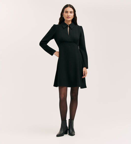 A woman with long dark hair and a neutral expression stands against a plain background wearing the Sabine Black Crepe Short Dress with long sleeves, a keyhole neckline, black tights, and ankle boots.