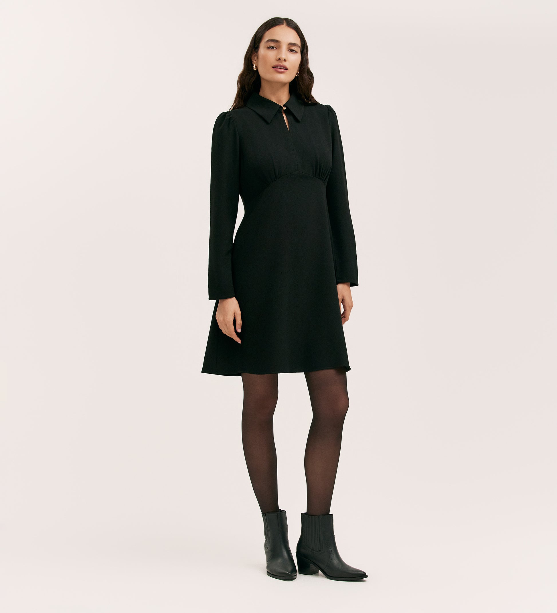 A woman with long, wavy hair and a neutral expression stands against a plain background wearing the Sabine Black Crepe Short Dress with long sleeves and a collar, paired with black tights and black ankle boots.