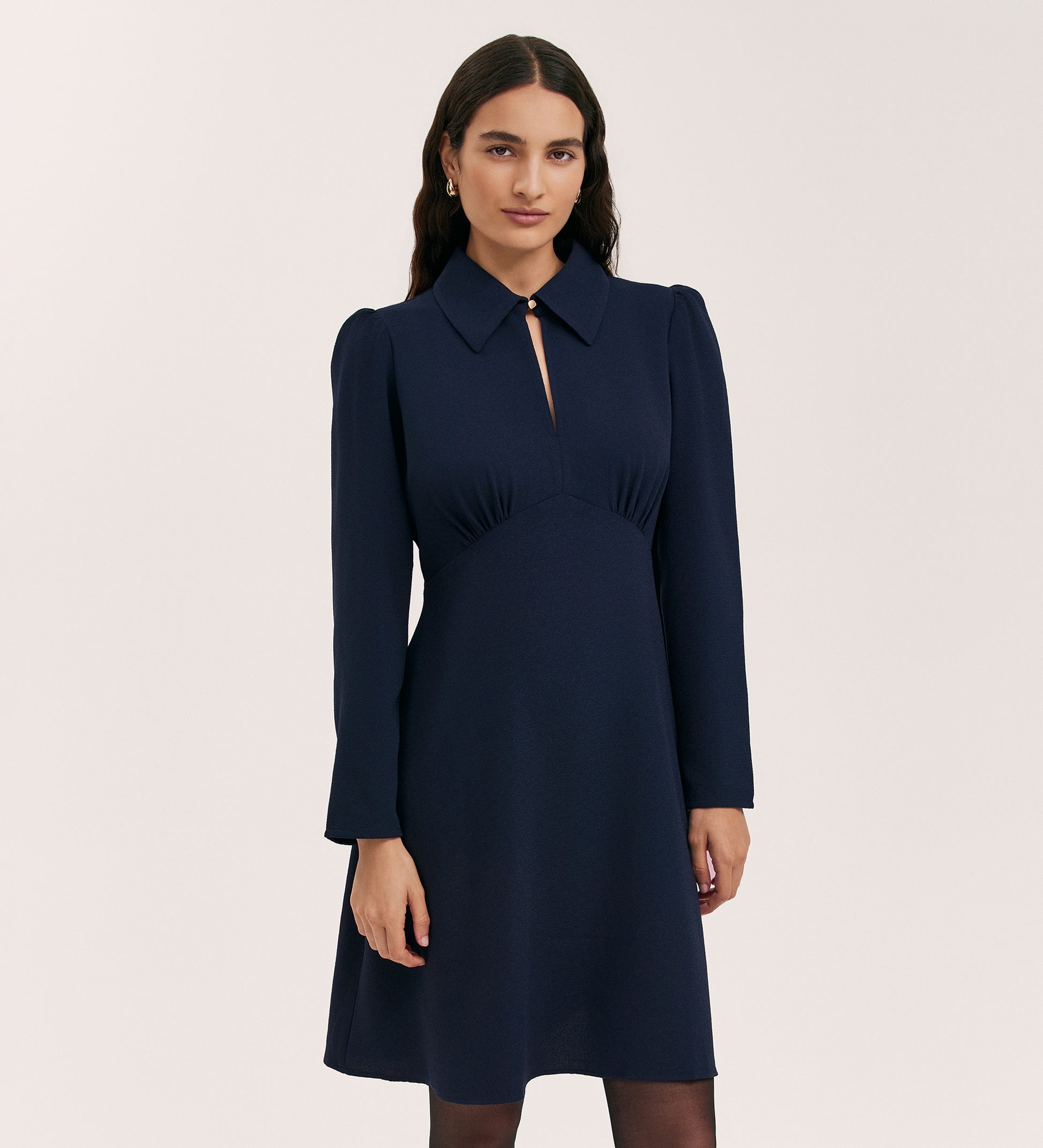 A woman with long, dark hair and a neutral expression models the Sabine Navy Crepe Short Dress, featuring long sleeves, a fitted cut, slight puff shoulders, and a pointed collar against a plain light background.