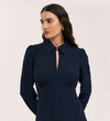 A woman with long dark hair wears the Sabine Navy Crepe Short Dress, featuring long sleeves, a deep collared V-neckline, subtle puffed shoulders, and a modern fitted silhouette, standing against a plain light background.