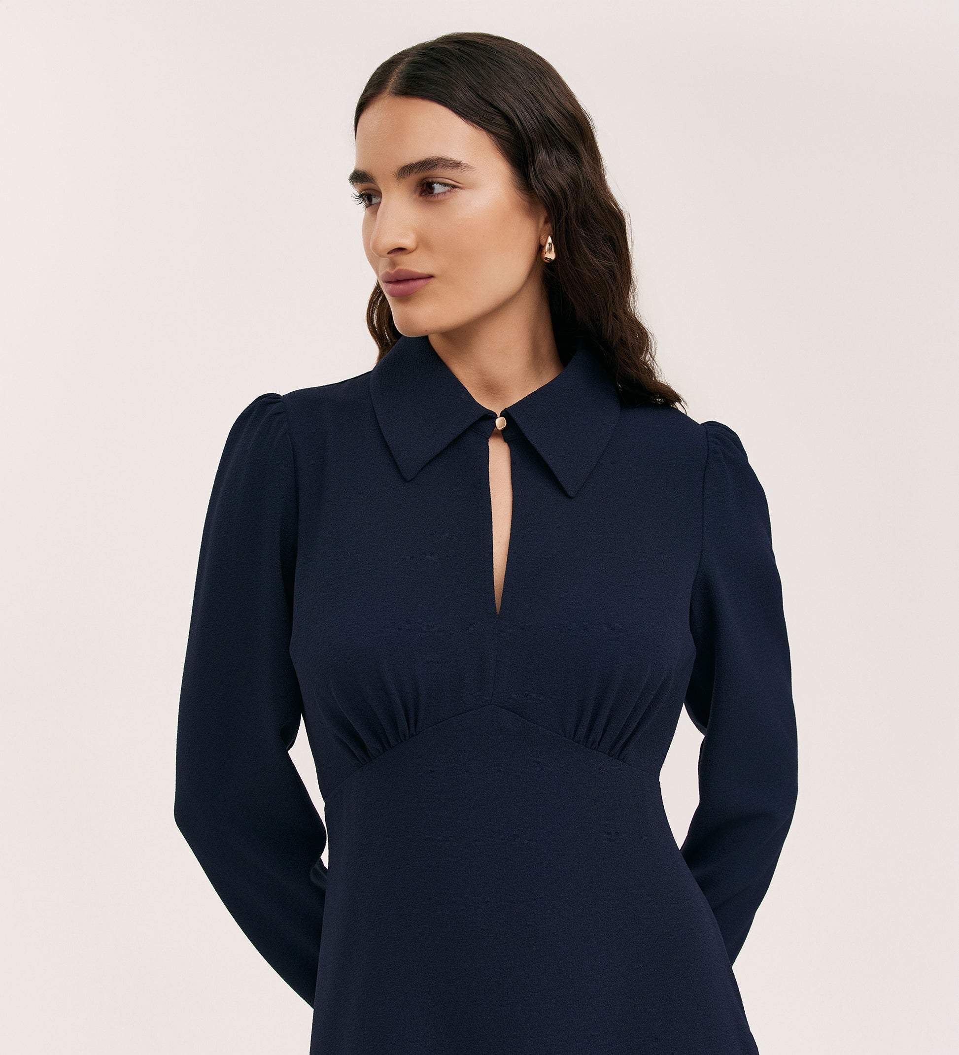 A woman with long dark hair wears the Sabine Navy Crepe Short Dress, featuring long sleeves, a deep collared V-neckline, subtle puffed shoulders, and a modern fitted silhouette, standing against a plain light background.