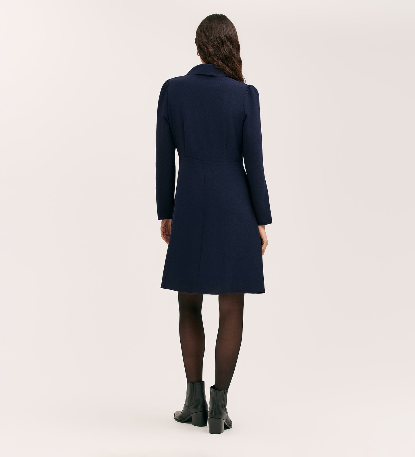 A woman faces away, wearing the Sabine Navy Crepe Short Dress with a fitted cut, styled with black tights and ankle boots. Her long hair is loose against a plain, light background.