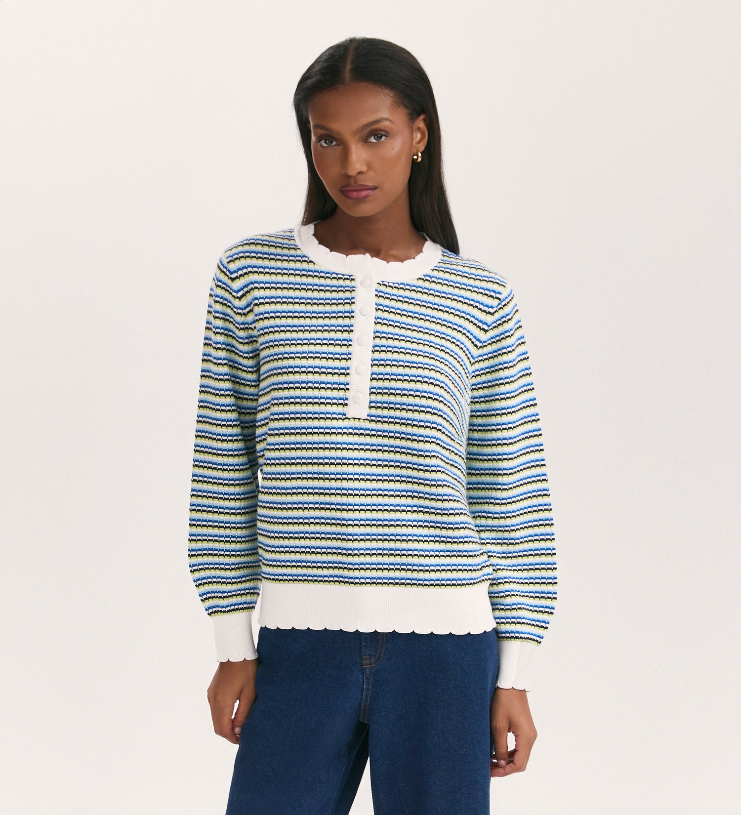 A woman with straight, dark hair wears the Sanne Blue Stripe Jumper—featuring long sleeves, stripes, scallop rib detail, and a partial button placket—with blue jeans against a plain white background.