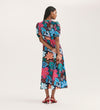 A woman with long dark hair wears the Sarah Blue Floral Crepe Midi Dress—featuring a colorful floral pattern, button-through front, and short sleeves—with red heeled sandals, set against a plain light background.
