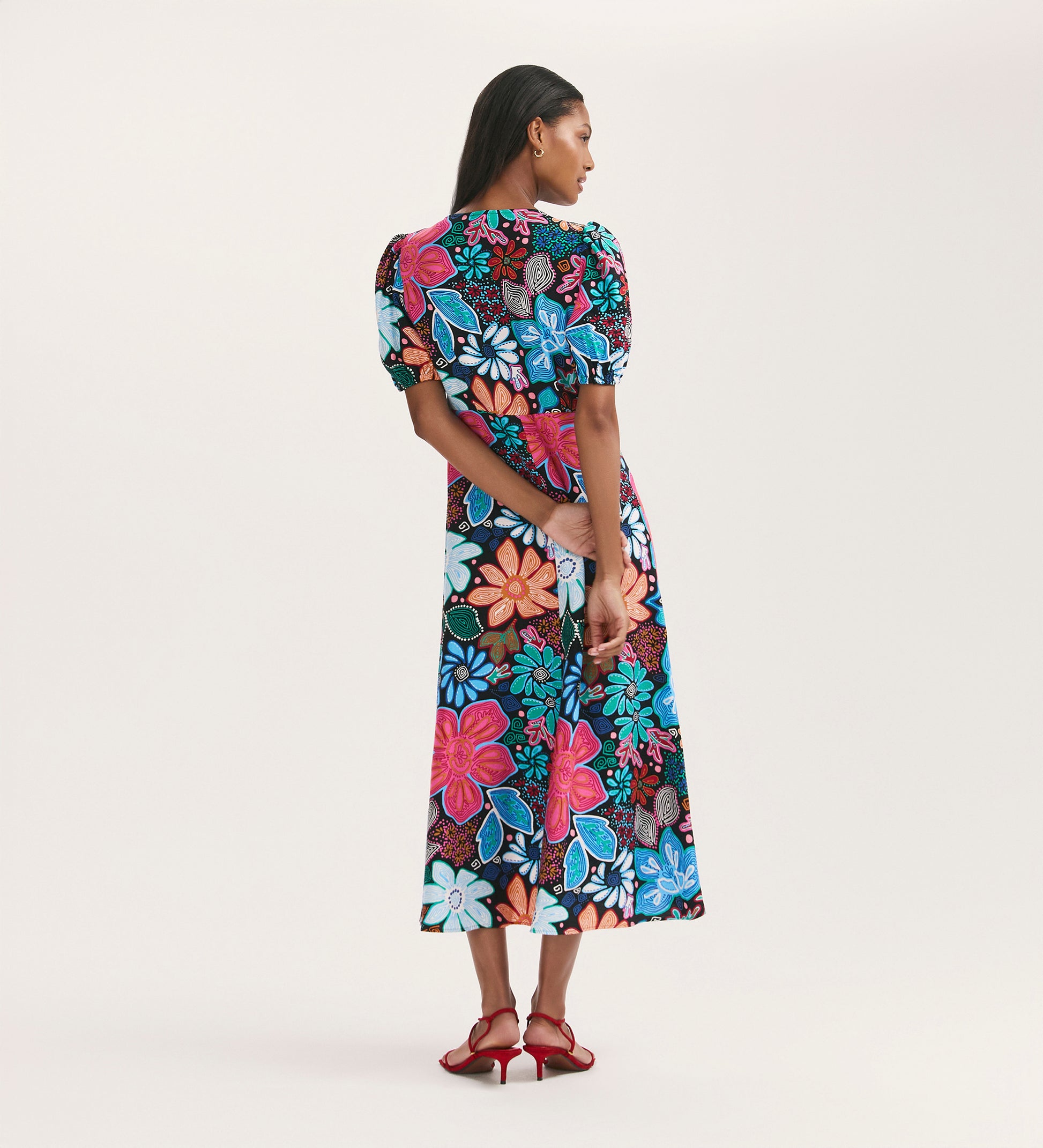 A woman with long dark hair wears the Sarah Blue Floral Crepe Midi Dress—featuring a colorful floral pattern, button-through front, and short sleeves—with red heeled sandals, set against a plain light background.