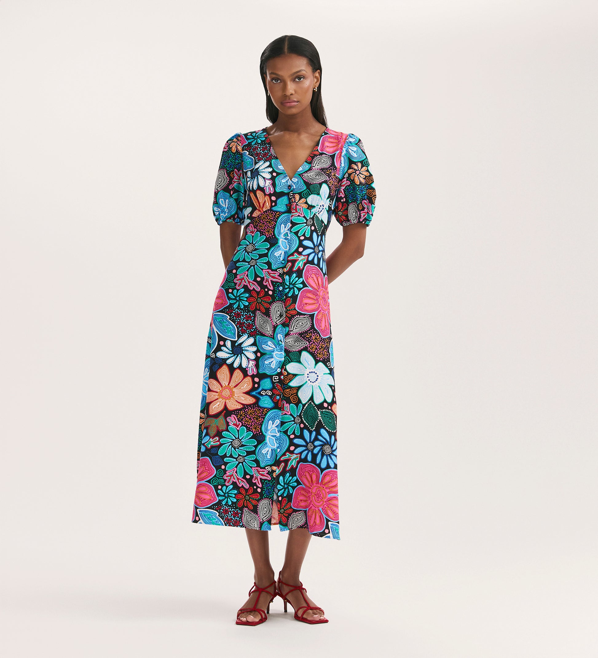 A woman wears the Sarah Blue Floral Crepe Midi Dress, featuring a V-neckline, short sleeves, and button-through front. Her straight hair and red strappy sandals complete the look against a plain light background.