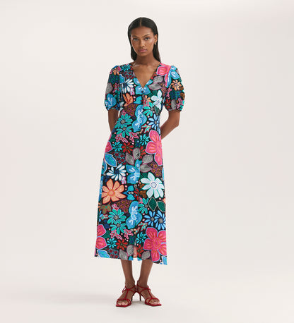 A woman wears the Sarah Blue Floral Crepe Midi Dress, featuring a V-neckline, short sleeves, and button-through front. Her straight hair and red strappy sandals complete the look against a plain light background.