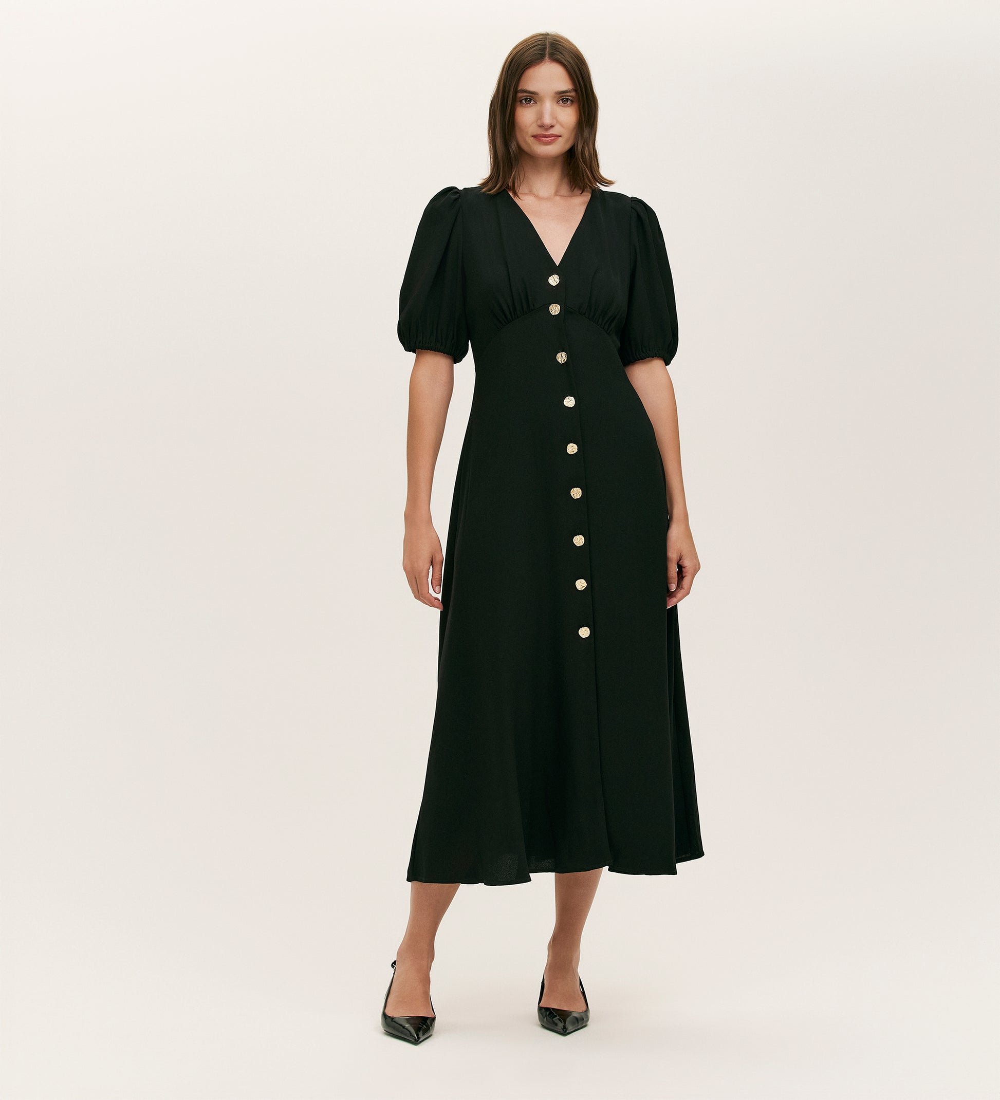 A woman models the Sarah Black Crepe Button Through Midi Tea Dress, featuring short puff sleeves, a V-neckline, and large gold front buttons. She pairs it with black pointed-toe heels against a plain background.
