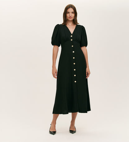 A woman models the Sarah Black Crepe Button Through Midi Tea Dress, featuring short puff sleeves, a V-neckline, and large gold front buttons. She pairs it with black pointed-toe heels against a plain background.