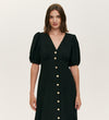A woman with straight brown hair models the Sarah Black Crepe Button Through Midi Tea Dress, showcasing short puff sleeves, a V-neckline, and gold front buttons against a plain light background.
