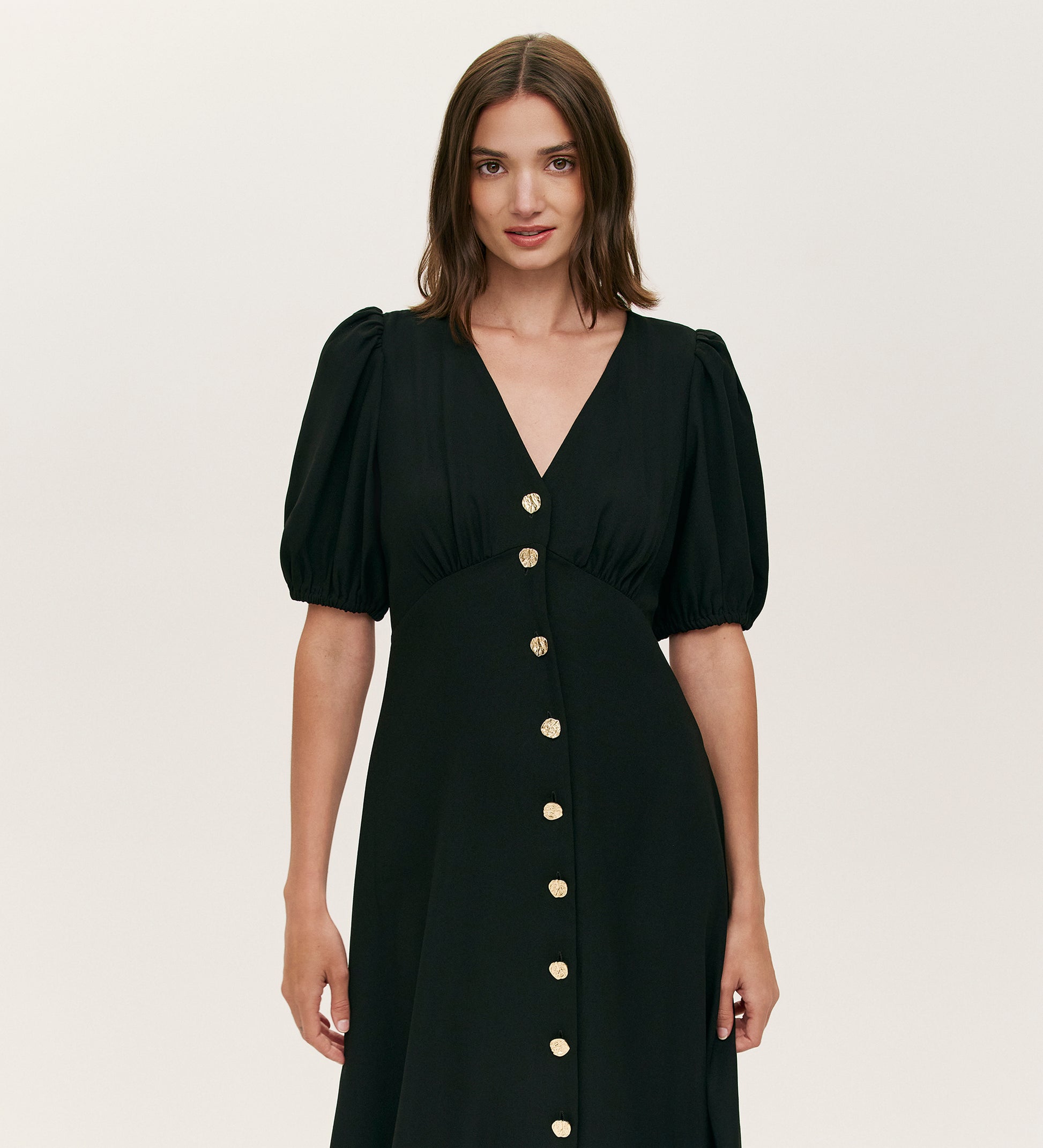 A woman with straight brown hair models the Sarah Black Crepe Button Through Midi Tea Dress, showcasing short puff sleeves, a V-neckline, and gold front buttons against a plain light background.