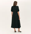 A woman faces away, wearing the Sarah Black Crepe Button Through Midi Tea Dress with short puff sleeves and a V-neckline, paired with black slingback heels, set against a plain light background.