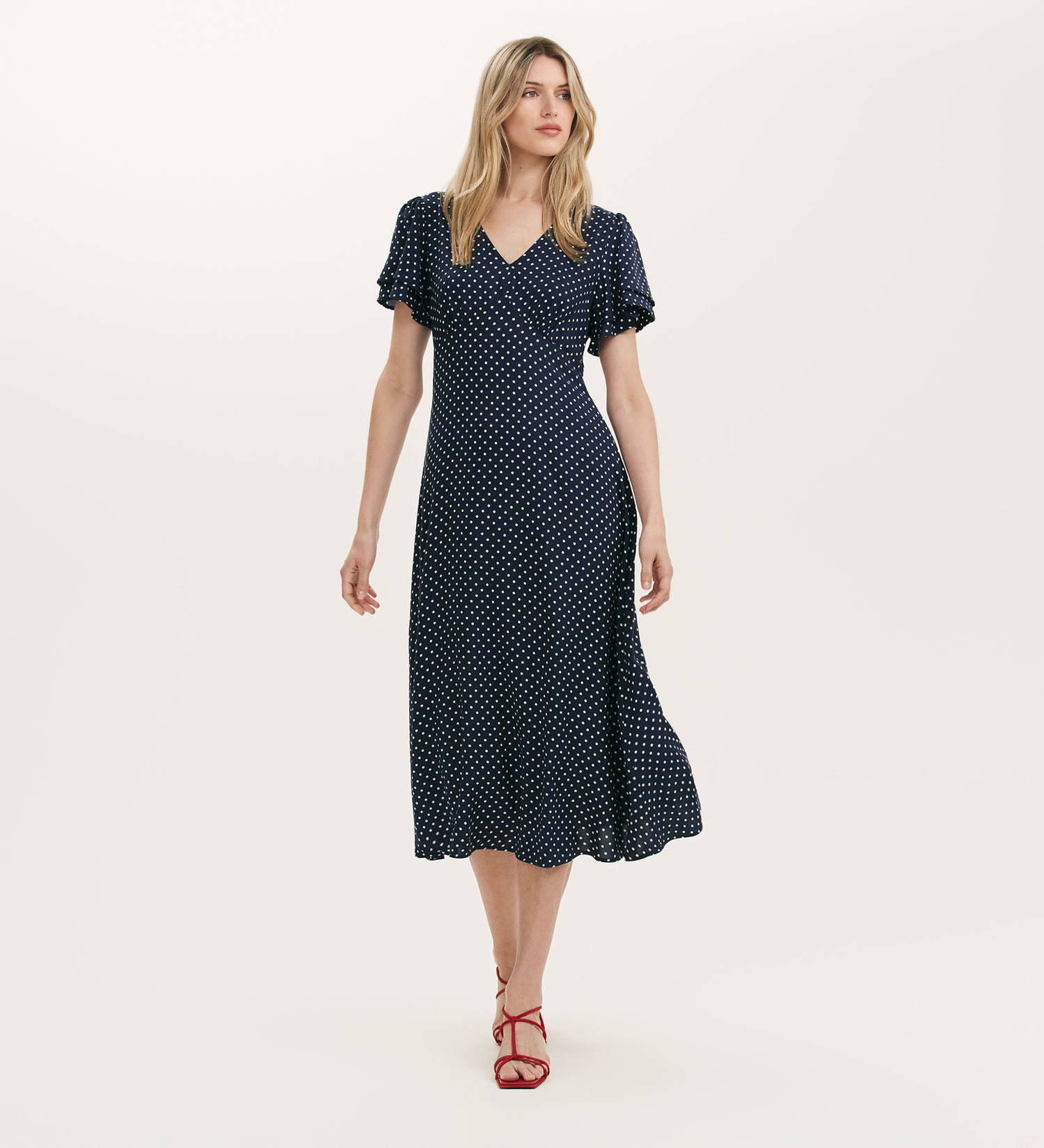 A woman wearing the Saskia Navy Polka Dot Midi Tea Dress with red strappy heels stands against a plain light background.