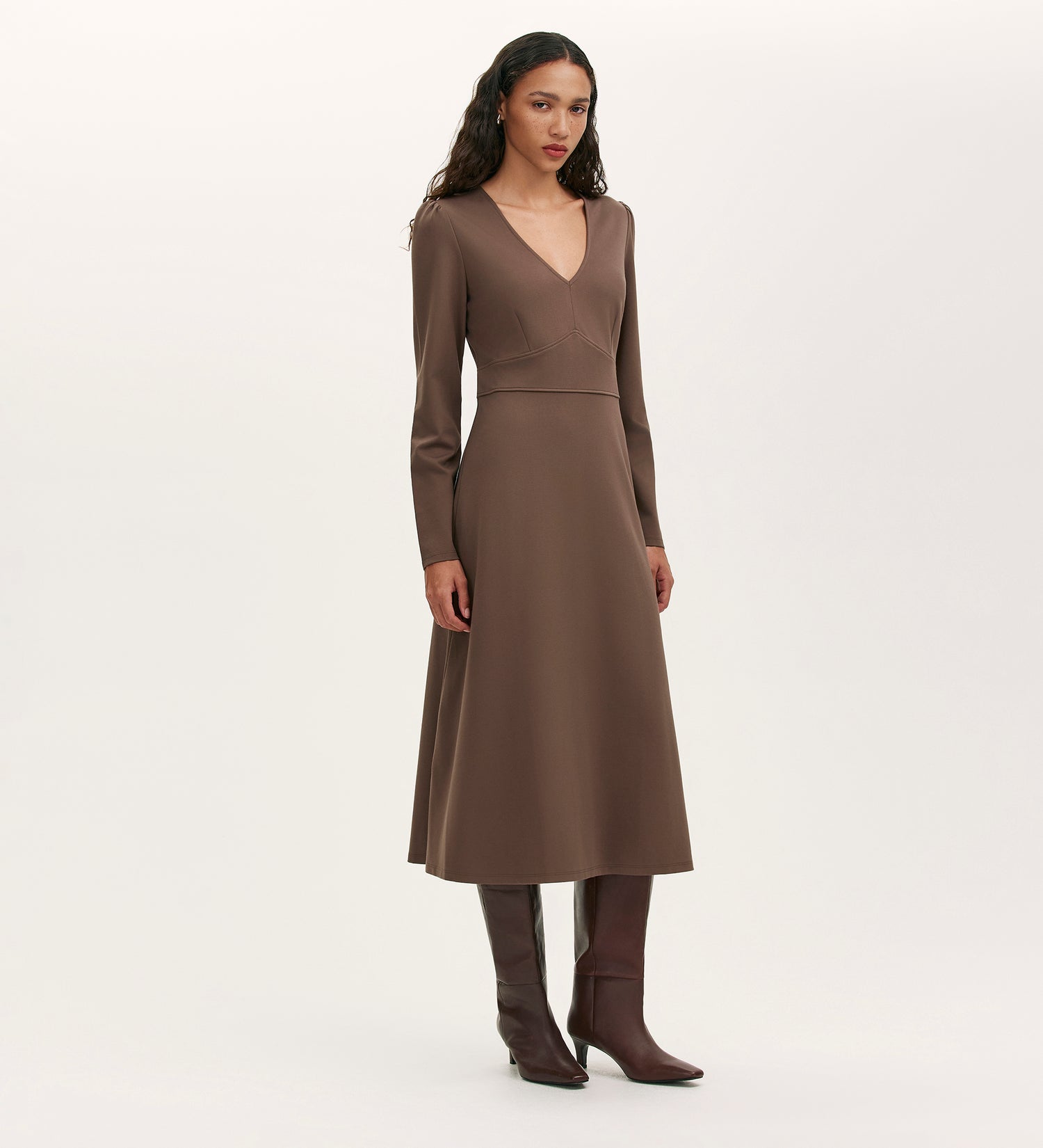 A woman stands against a plain, light background wearing the Seraphina Mole Ponte Jersey Long Sleeve Midi Dress, featuring a V-neckline, paired with brown knee-high boots.