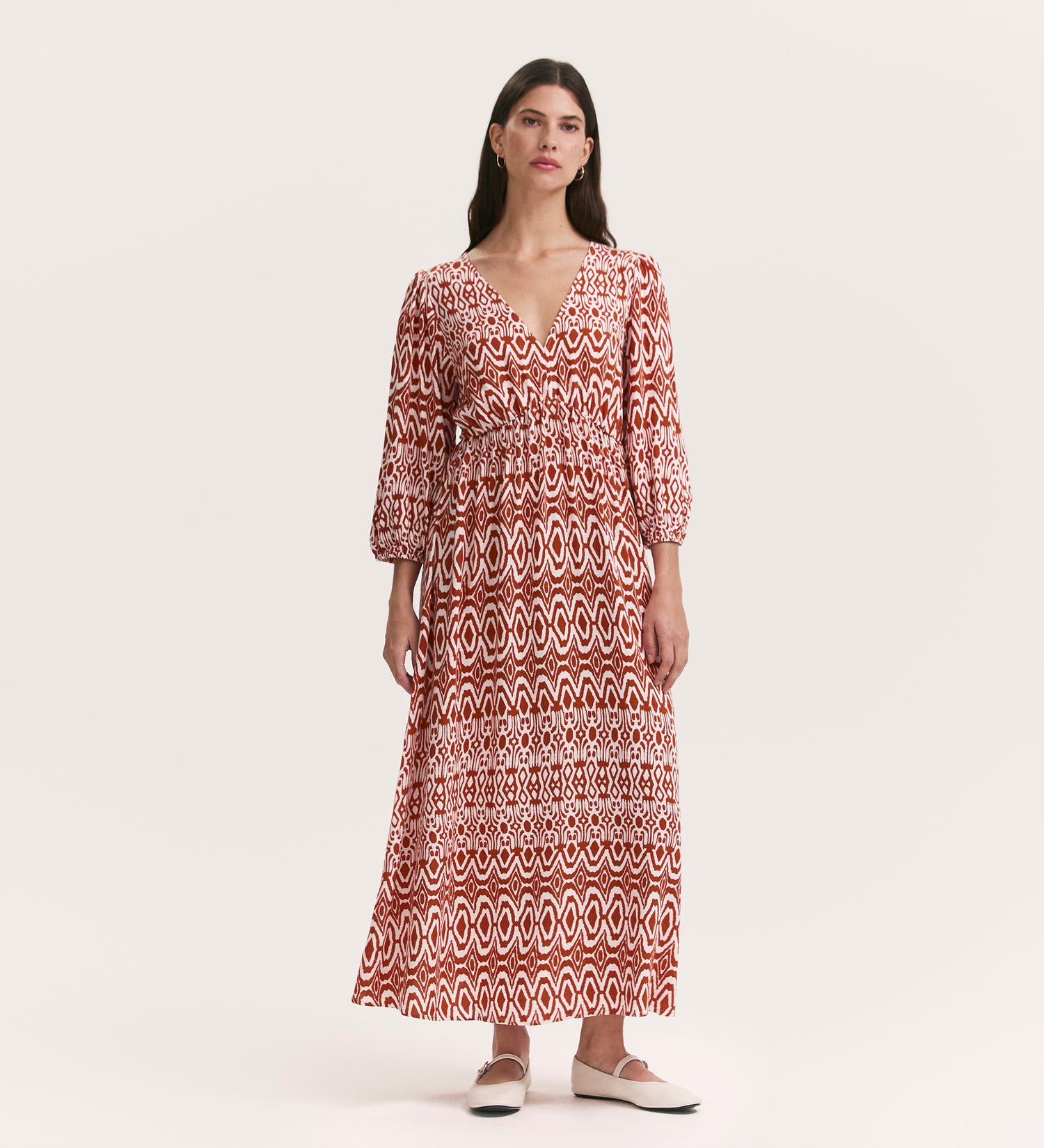 A woman models the Serena Brown Tribal Crepe Midi Smock Dress with a deep V-neck, three-quarter sleeves, and a flowy fit. The dress features a bold tribal print. She pairs it with white flats and poses against a plain background.