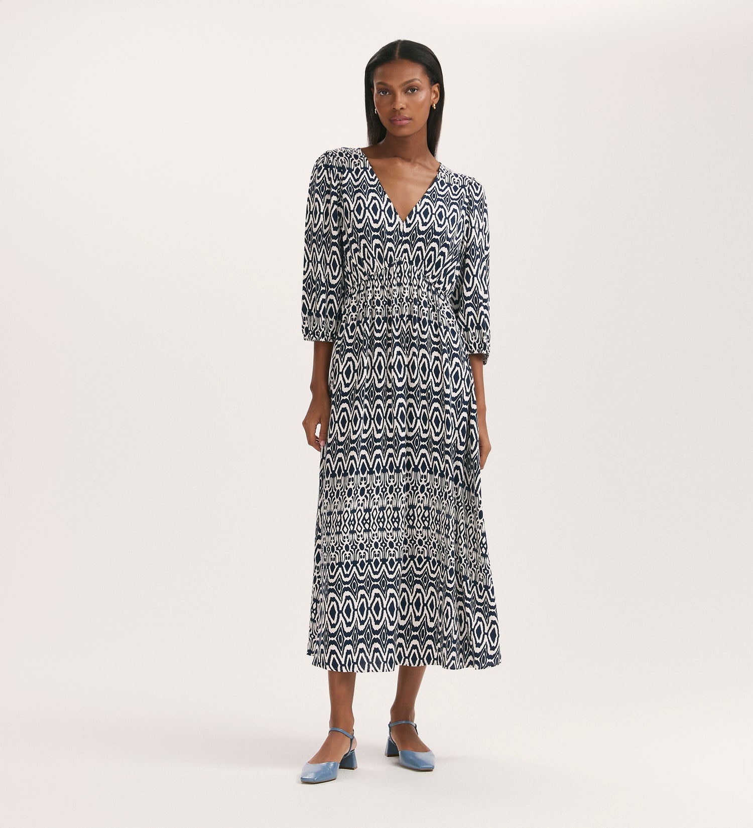 A woman with straight dark hair stands against a plain background, wearing the Serena Navy Tribal Crepe Midi Smock Dress featuring a fit and flare silhouette, three-quarter sleeves, a V-neckline, and paired with blue low-heeled shoes.