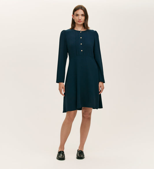 A woman stands against a plain background wearing the Shauna Navy Crepe Short Dress, a long-sleeve, knee-length piece with gold button details on the front, paired with black loafers.