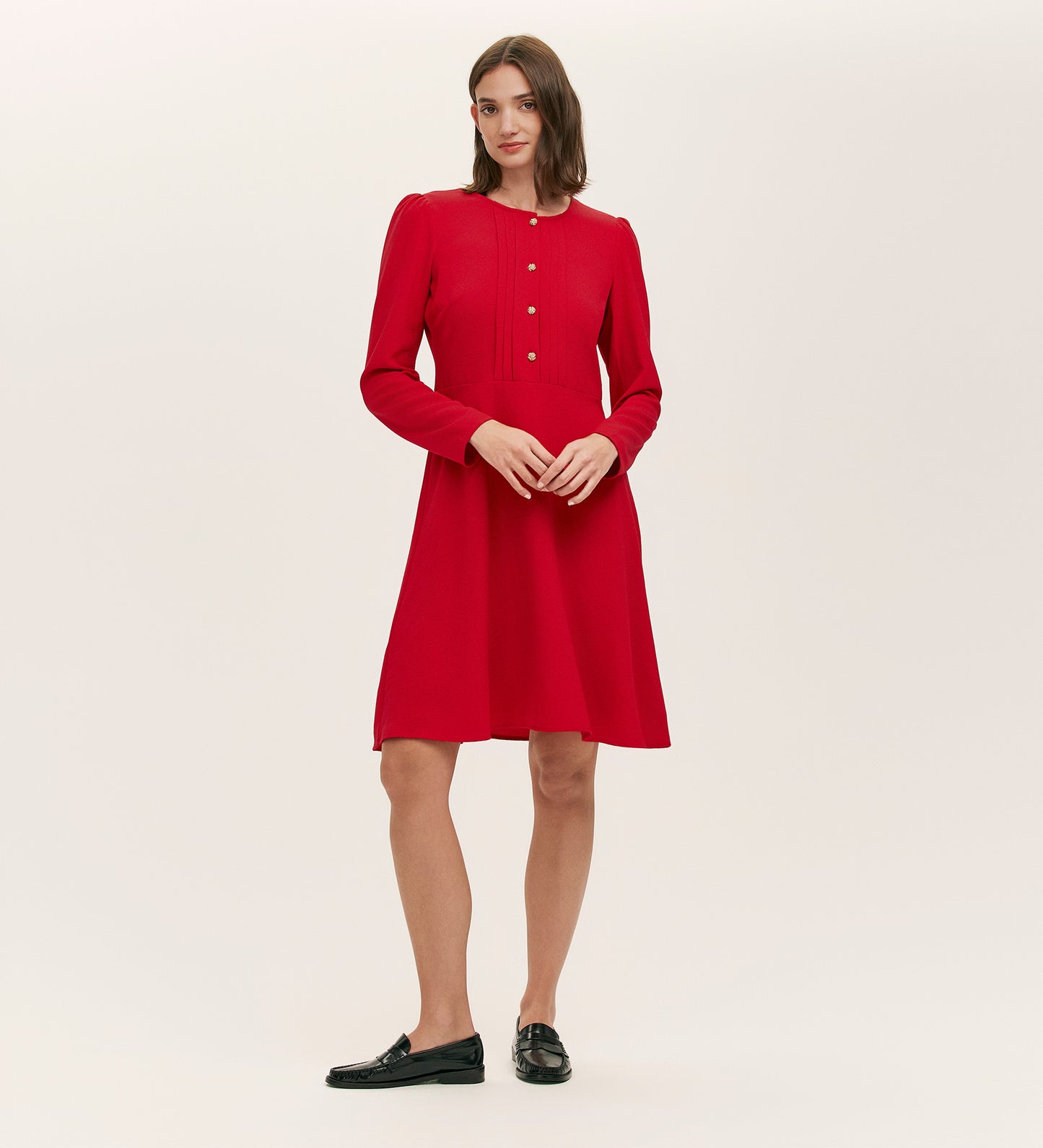 A woman poses confidently in the Shauna Red Crepe Short Dress, featuring signature crepe fabric and gold button detailing. She pairs it with black loafers, her straight shoulder-length brown hair complementing the chic look.