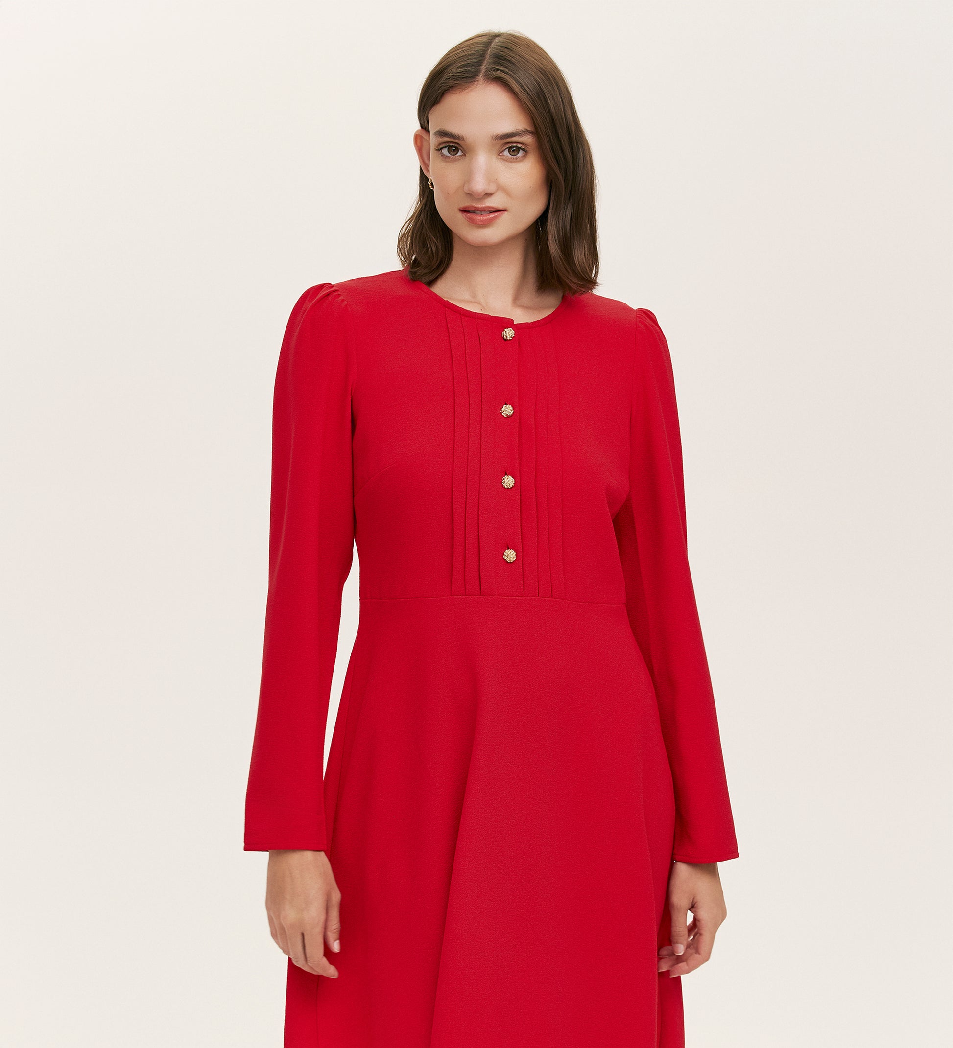 A woman with straight brown hair wears the Shauna Red Crepe Short Dress, featuring long sleeves, gold button details, and subtle bodice pleats, standing against a plain light background.