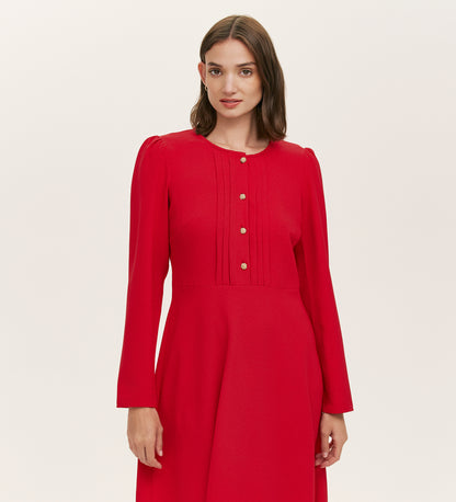 A woman with straight brown hair wears the Shauna Red Crepe Short Dress, featuring long sleeves, gold button details, and subtle bodice pleats, standing against a plain light background.