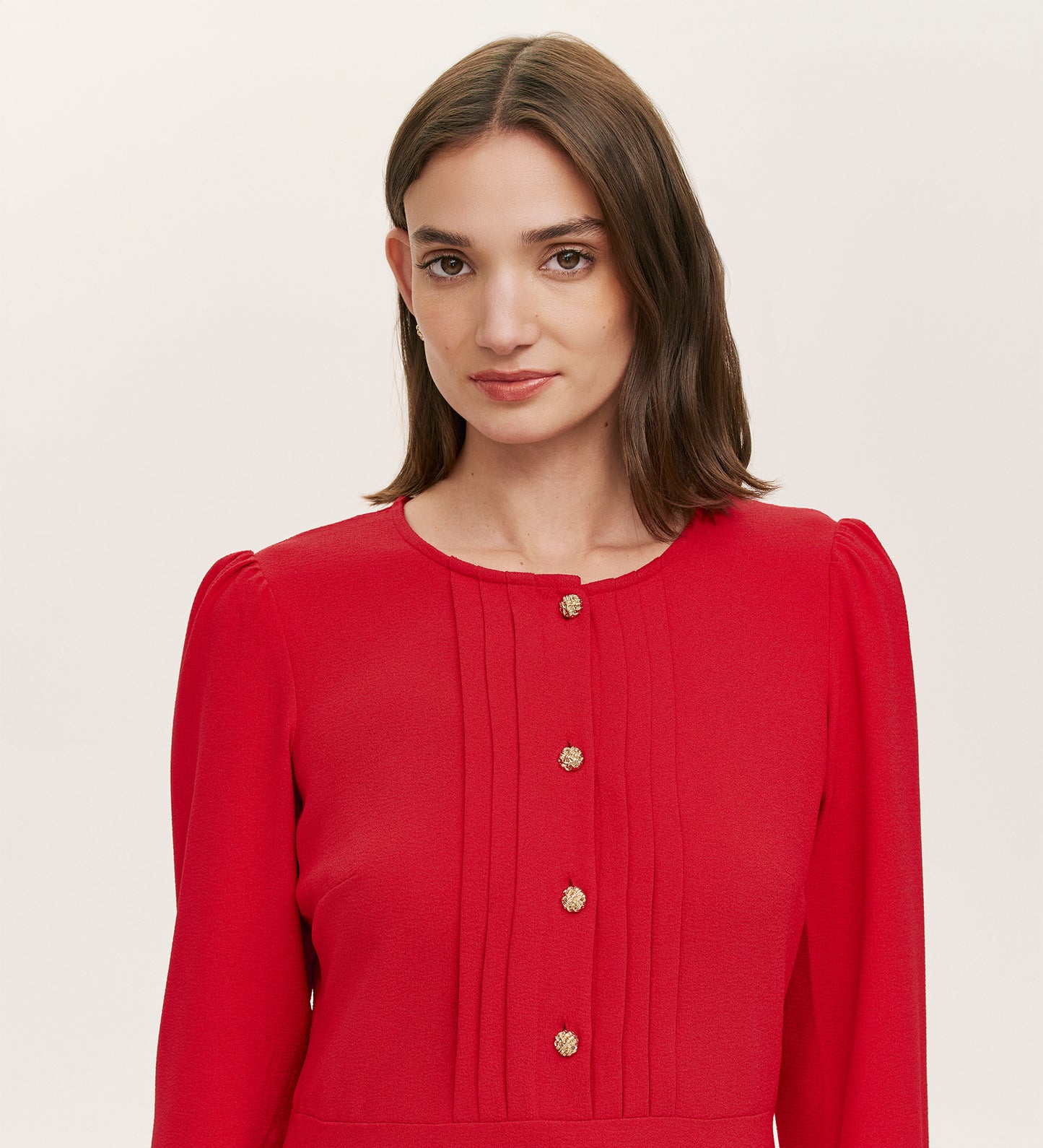 A woman with straight brown hair wears the Shauna Red Crepe Short Dress, a chic mini in bright red crepe with gold button details and front pleated accents, standing against a plain light background.