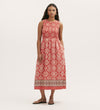 A woman with long, wavy hair wears the Skylar Coral Mosaic Midi Dress—a sleeveless midi dress with a shirred waist and border print hem in white geometric patterns on red—paired with sandals against a plain light background.