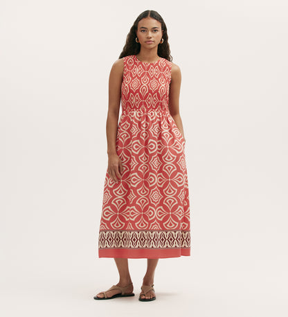 A woman with long, wavy hair wears the Skylar Coral Mosaic Midi Dress—a sleeveless midi dress with a shirred waist and border print hem in white geometric patterns on red—paired with sandals against a plain light background.