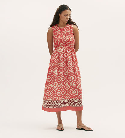 A woman with long curly hair stands with her hands behind her back, looking down, wearing the Skylar Coral Mosaic Midi Dress—sleeveless with a shirred waist and border print hem—and flat sandals.
