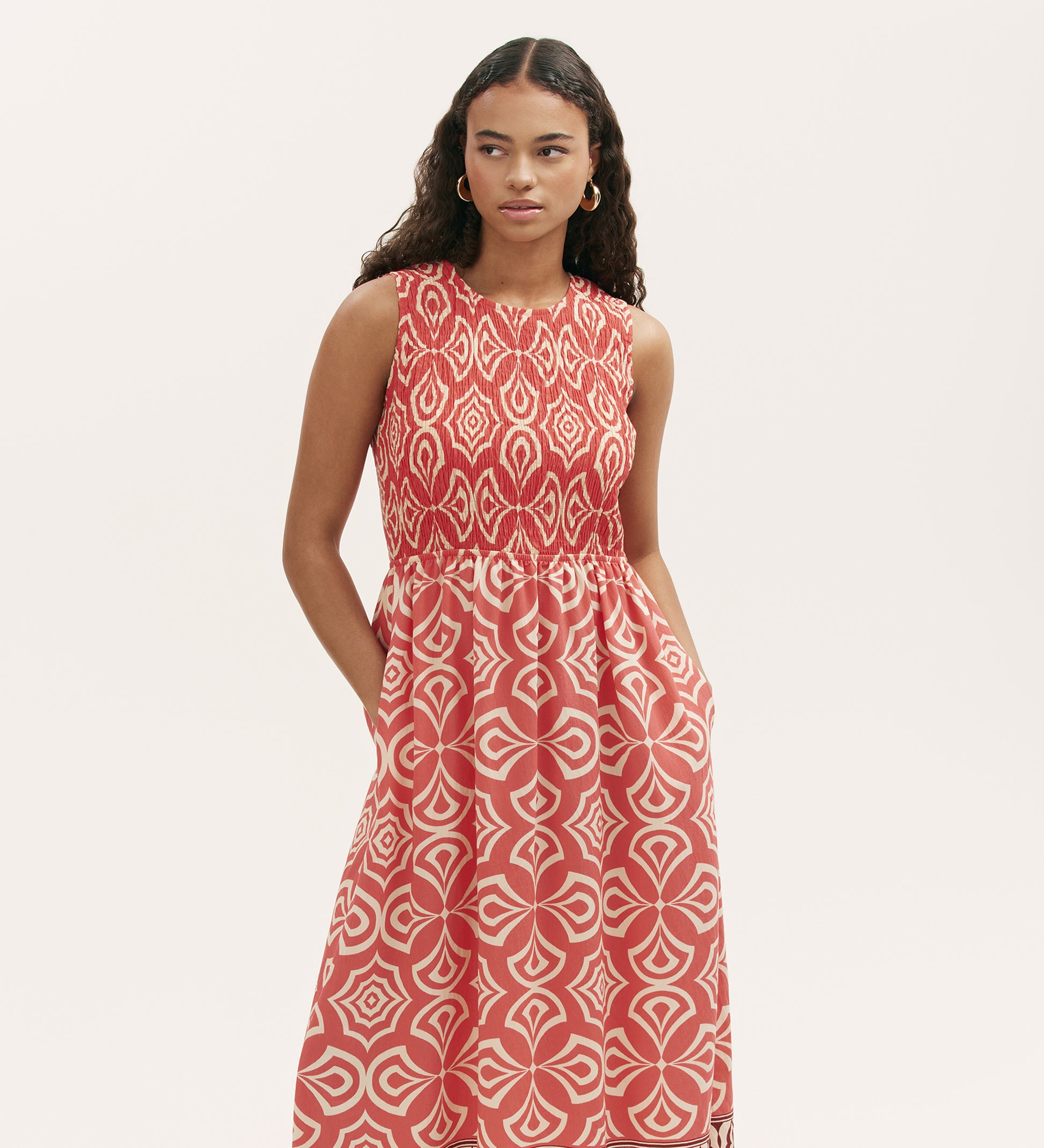 A woman with long curly hair wears the Skylar Coral Mosaic Midi Dress, featuring a sleeveless design, geometric pattern, shirred waist, and border print hem, standing against a plain white background.