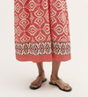 A person wearing the Skylar Coral Mosaic Midi Dress, featuring geometric patterns and a border print hem, stands in tan sandals against a plain background. Only the lower legs and feet are visible.