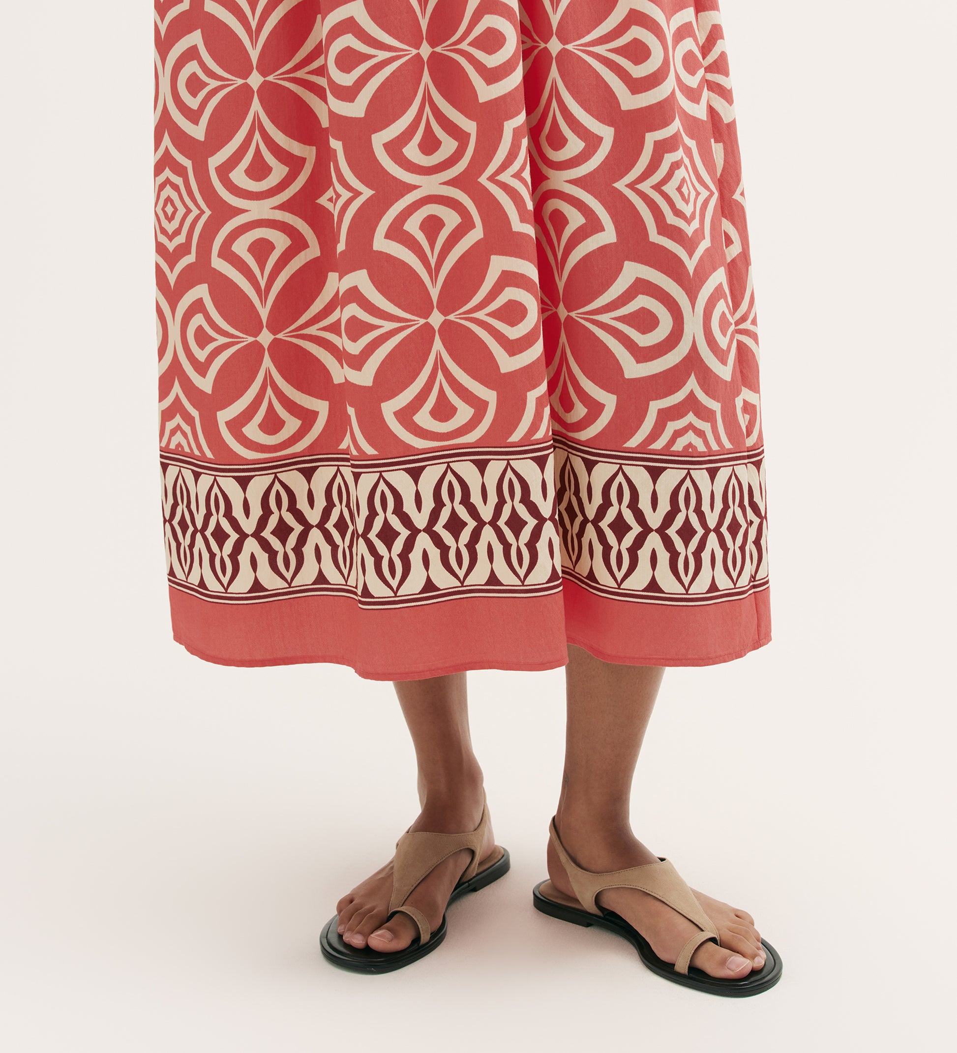 A person wearing the Skylar Coral Mosaic Midi Dress, featuring geometric patterns and a border print hem, stands in tan sandals against a plain background. Only the lower legs and feet are visible.