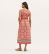A woman stands with her back to the camera, wearing the Skylar Coral Mosaic Midi Dress—sleeveless, featuring red and white geometric patterns, a shirred waist, and a border print hem—paired with brown sandals against a light background.