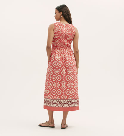 A woman stands with her back to the camera, wearing the Skylar Coral Mosaic Midi Dress—sleeveless, featuring red and white geometric patterns, a shirred waist, and a border print hem—paired with brown sandals against a light background.