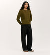 A woman with long, wavy dark hair wears the Valerie Khaki Knitted Jumper styled with loose black trousers featuring button details, standing against a plain light background.