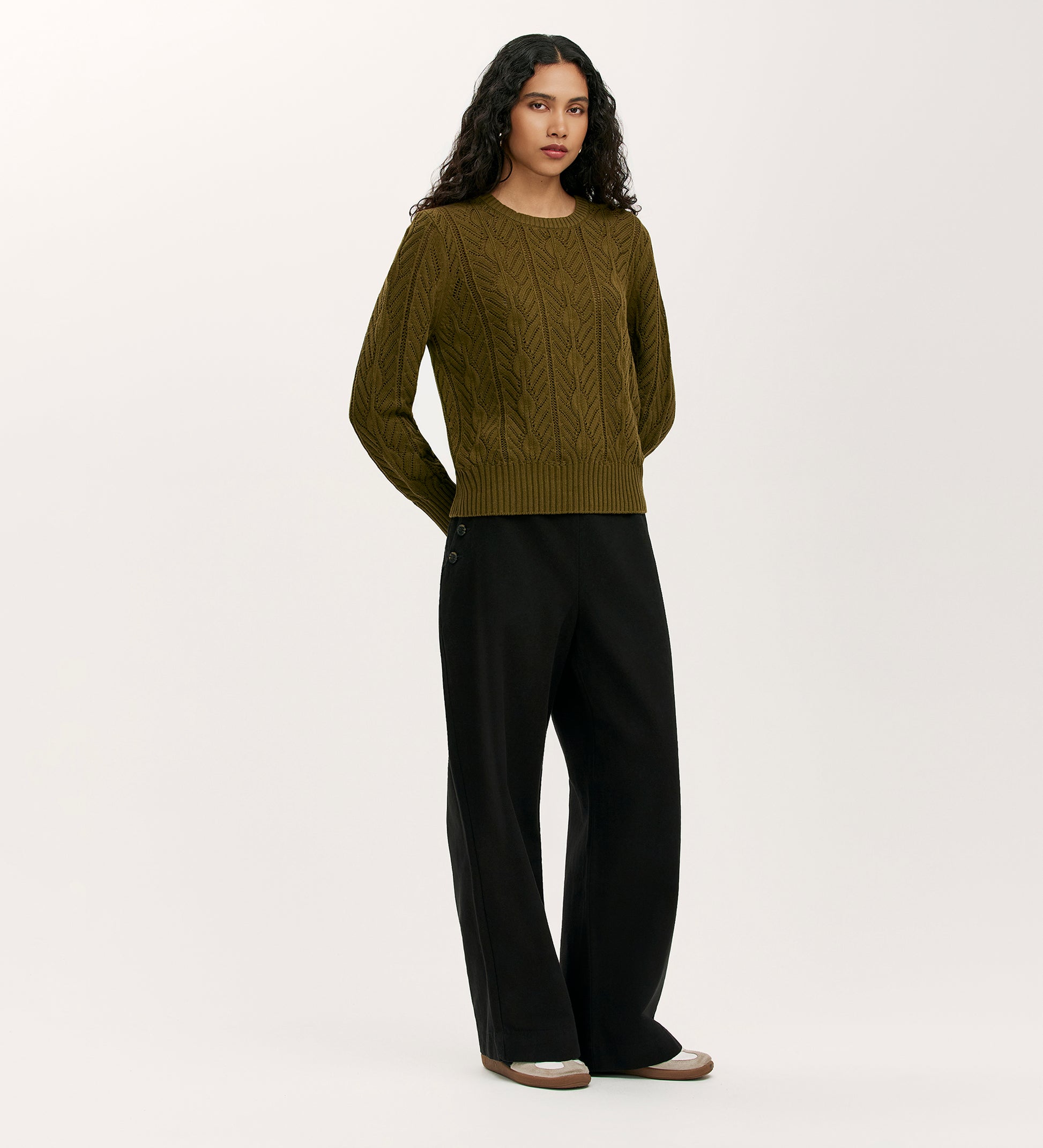 A woman with long, wavy dark hair wears the Valerie Khaki Knitted Jumper styled with loose black trousers featuring button details, standing against a plain light background.