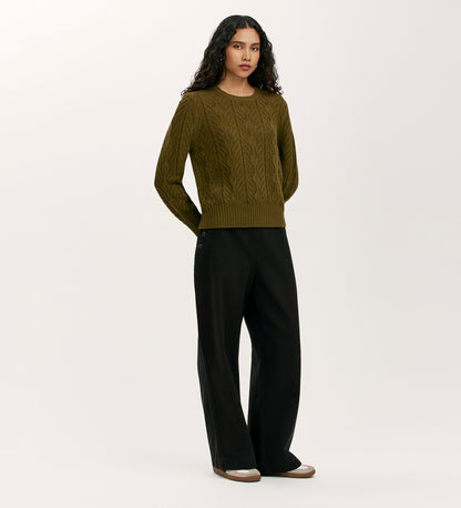 A woman with long, wavy dark hair wears the Valerie Khaki Knitted Jumper styled with loose black trousers featuring button details, standing against a plain light background.