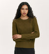 A woman with long curly hair stands with her arms crossed against a plain light background, wearing the Valerie Khaki Knitted Jumper—a stylish wardrobe essential.