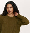 A woman with long curly hair and gold hoop earrings wears the relaxed-fit Valerie Khaki Knitted Jumper, looking to the side and touching her neck against a plain light background.
