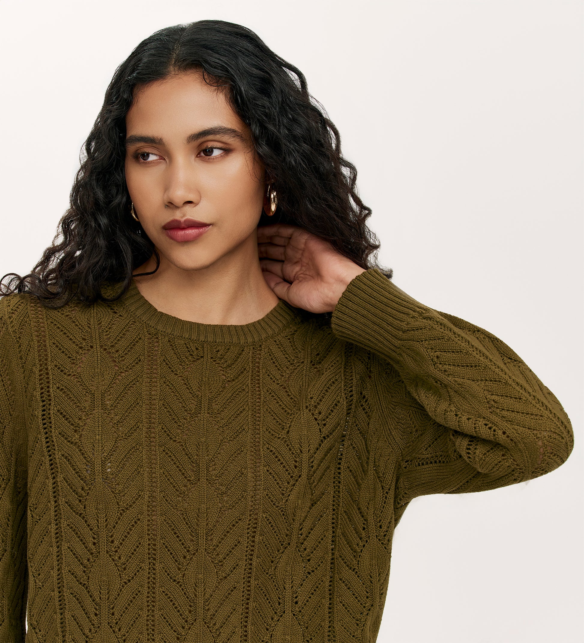 A woman with long curly hair and gold hoop earrings wears the relaxed-fit Valerie Khaki Knitted Jumper, looking to the side and touching her neck against a plain light background.