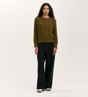 A woman with long curly hair models the Valerie Khaki Knitted Jumper, styled with wide-leg black pants and beige shoes, creating a relaxed wardrobe essential look against a plain light background.