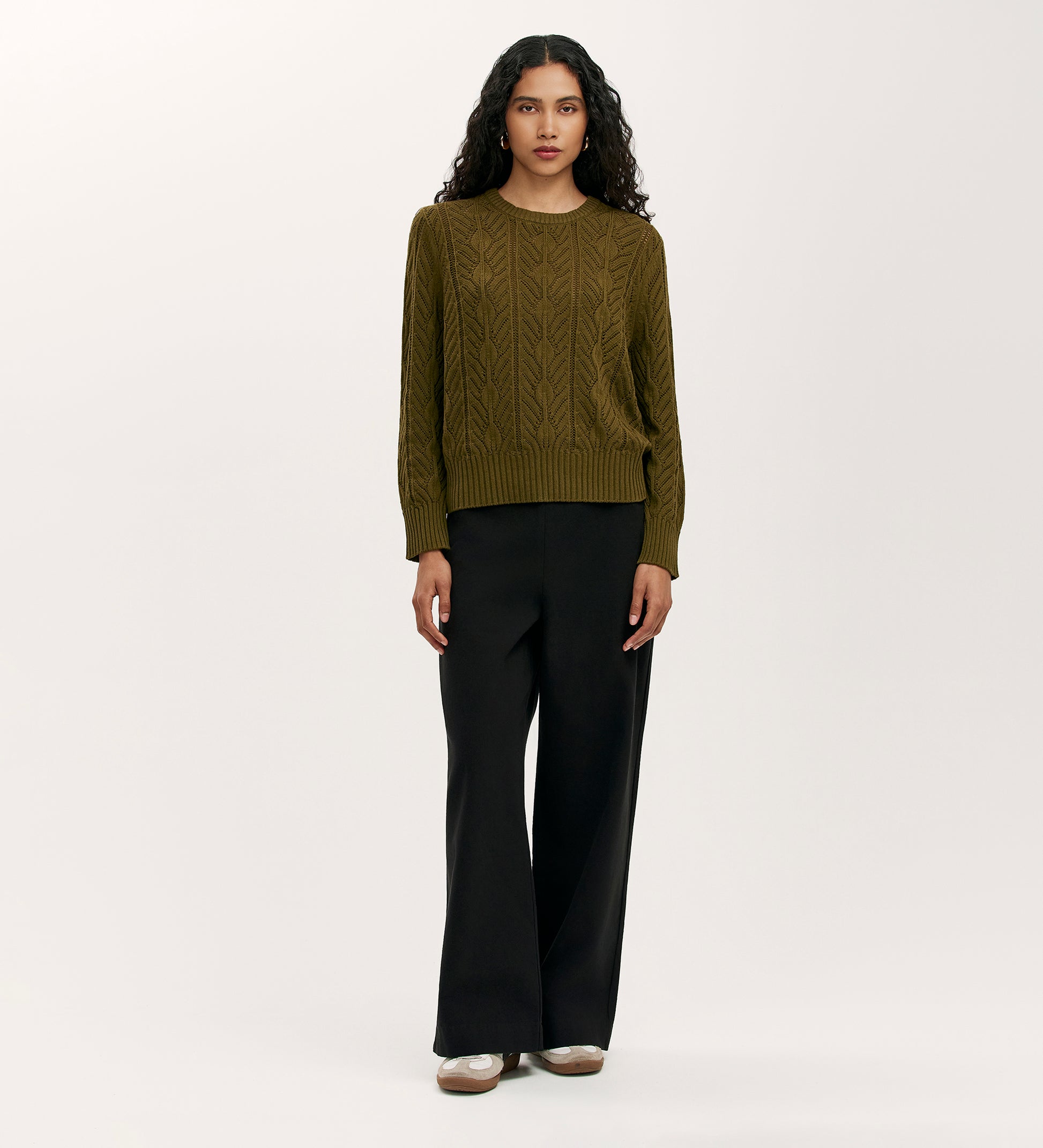 A woman with long curly hair models the Valerie Khaki Knitted Jumper, styled with wide-leg black pants and beige shoes, creating a relaxed wardrobe essential look against a plain light background.