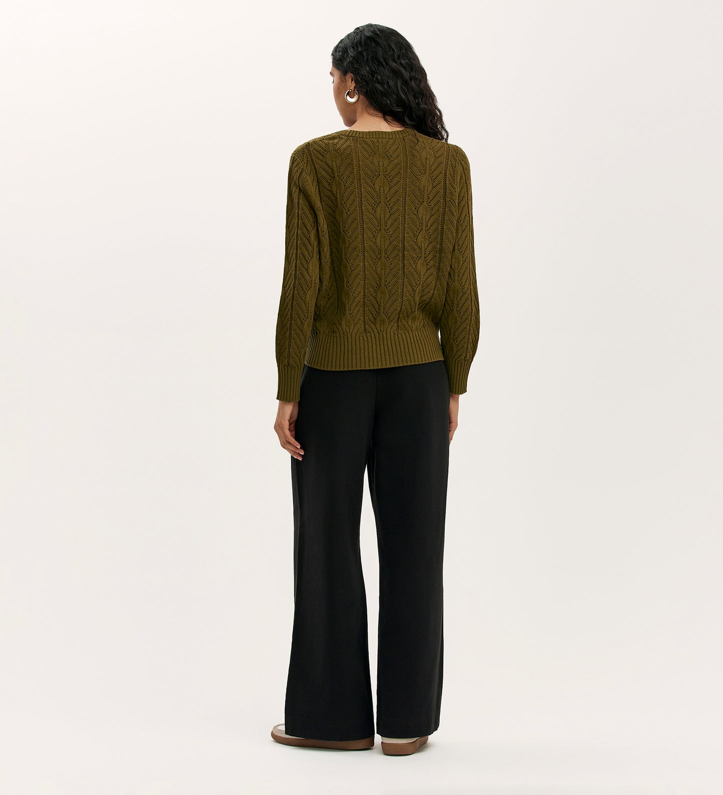 A person with long, wavy hair faces away, wearing the Valerie Khaki Knitted Jumper, wide-leg black pants, and brown shoes against a plain light background.