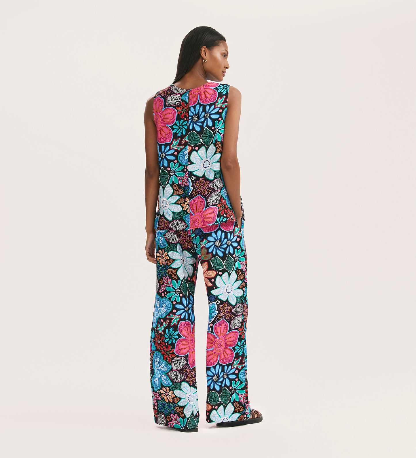 A woman with long hair stands against a plain background, wearing Eden Blue Floral Crepe Wide Leg Trousers featuring a colorful, bold floral print in blue, pink, red, and white.