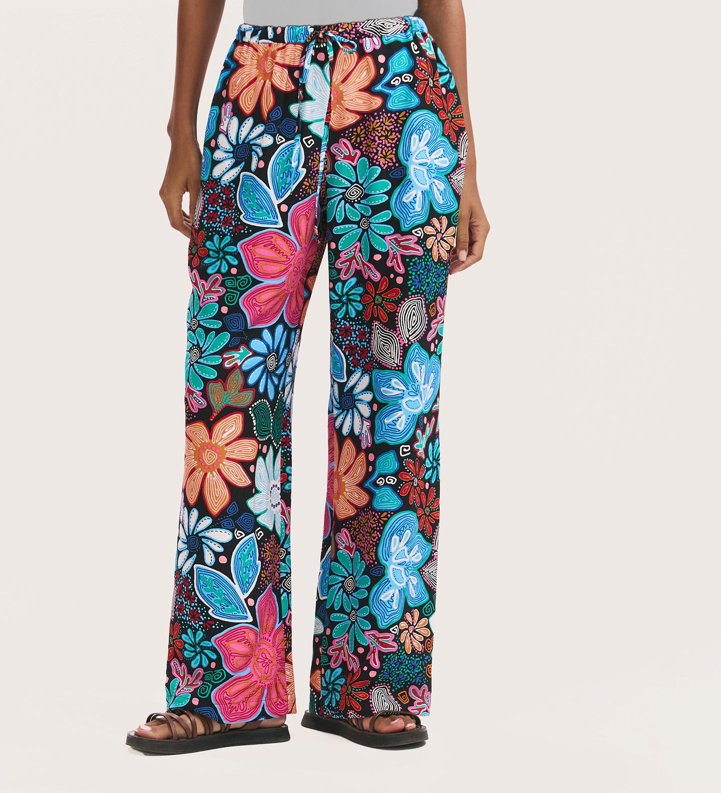 Wearing the Eden Blue Floral Crepe Wide Leg Trousers with a bold blue, pink, orange, and teal floral pattern, elasticated waistband, and side pockets, this person stands hands at sides in black sandals.