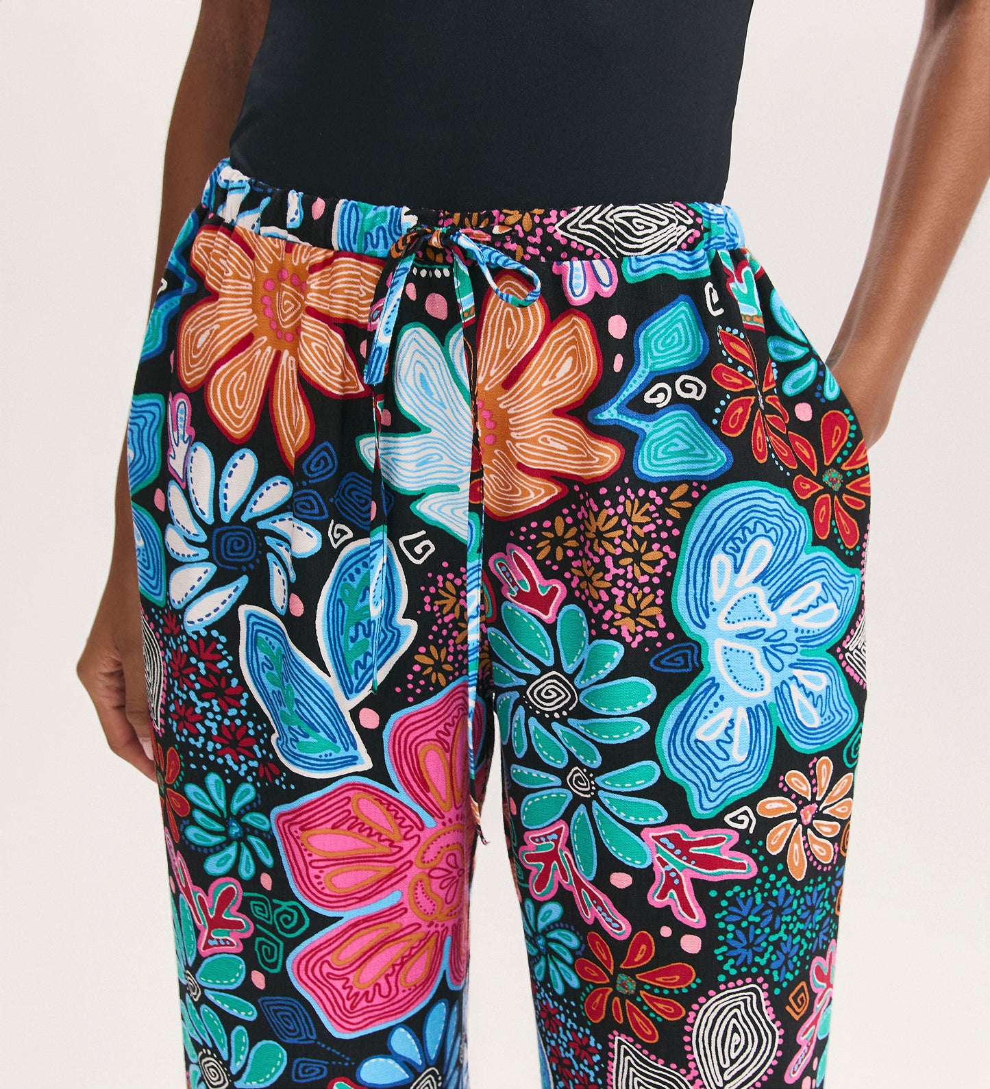 Seen from the waist down, a person wears Eden Blue Floral Crepe Wide Leg Trousers—colorful, floral-patterned with an elastic black waistband, drawstring tie, and side pockets—paired with a black top. Only the lower torso and arms are visible.
