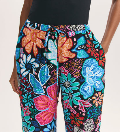 Seen from the waist down, a person wears Eden Blue Floral Crepe Wide Leg Trousers—colorful, floral-patterned with an elastic black waistband, drawstring tie, and side pockets—paired with a black top. Only the lower torso and arms are visible.