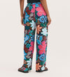 A person wears Eden Blue Floral Crepe Wide Leg Trousers, featuring a vibrant multicolored floral print in blue, pink, red, and white with an elasticated waistband, standing against a plain light background.